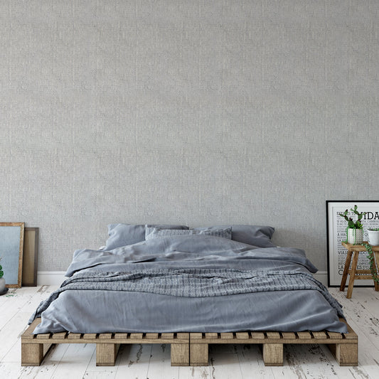 A bedroom with a bed on a pallette frame with photos either side featuring 'Alina' which is a grey wallpaper featuring a soft wool fabric-effect texture with a woven appearance and subtle tonal variation in a minimalist repeating design.