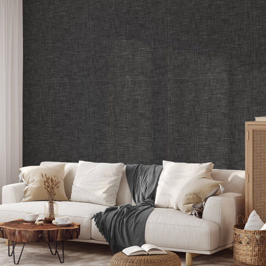A living room with light coloured sofa and rattan furnitire featuring 'Ashlyn' which is a dark grey wallpaper featuring a subtle linen fabric-effect texture with a woven appearance and soft tonal variation in a minimalist design.