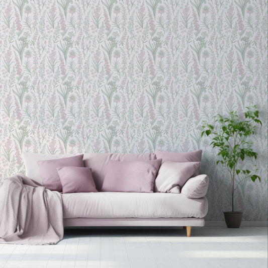 A living room with a dusty pink velour sofa scattered with pillows and blankets, next to a free standing leafy green plant showing 'Esther' which is a Victorian-inspired wallpaper featuring delicate pink foxglove flowers, green leaves and botanical stems on a soft pale pink background in a repeating floral pattern.