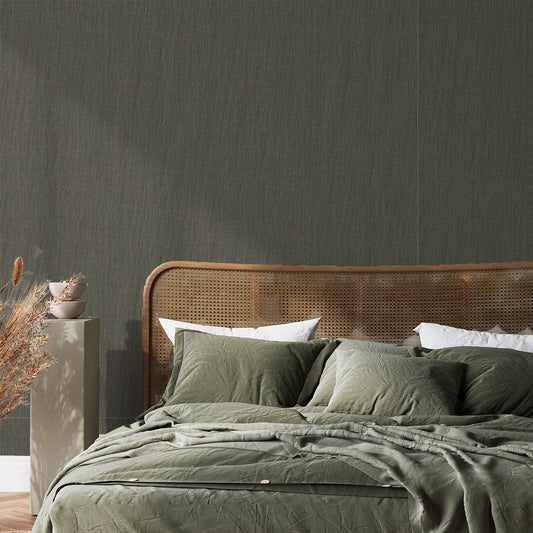 A modern bedroom with a rattan bedframe and dark green bedding, showing 'Hazel' which is a dark green wallpaper featuring a subtle fabric-effect texture with a woven pattern and grey undertones for a soft, sophisticated finish.
