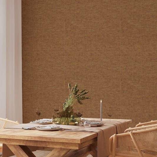 A dining room with rattan style furniture showing 'Olive' which is a brown wallpaper featuring a subtle grass cloth texture with woven fibres and natural tonal variations in a repeating textured pattern.