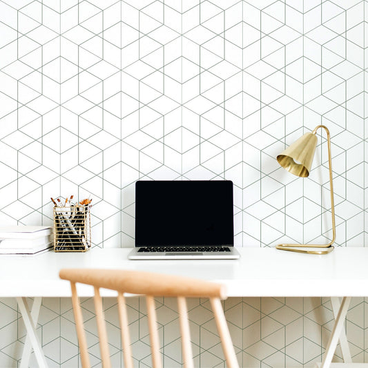 An office room with a white desk with a laptop, lamp and stationary on top featuring 'Rosamund' which is a dark green geometric wallpaper featuring overlapping diamond outlines and fine linear detailing in a modern repeating pattern.