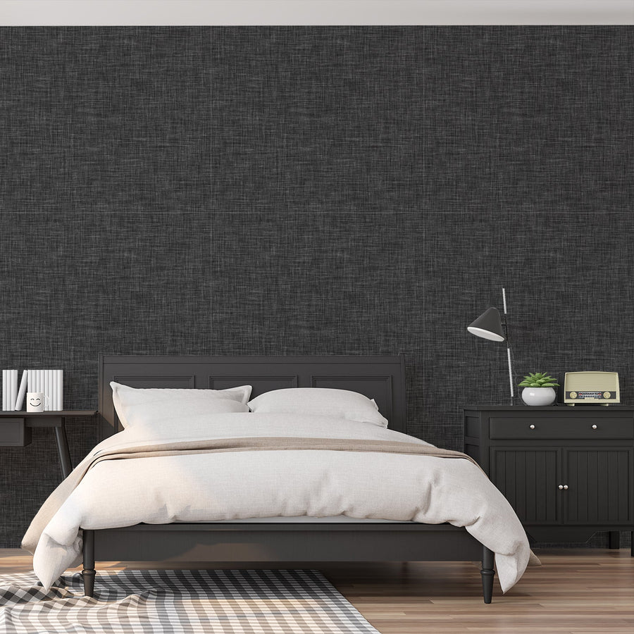 A bedroom with dark bed frame and side table featuring 'Ashlyn' which is a dark grey wallpaper featuring a subtle linen fabric-effect texture with a woven appearance and soft tonal variation in a minimalist design.