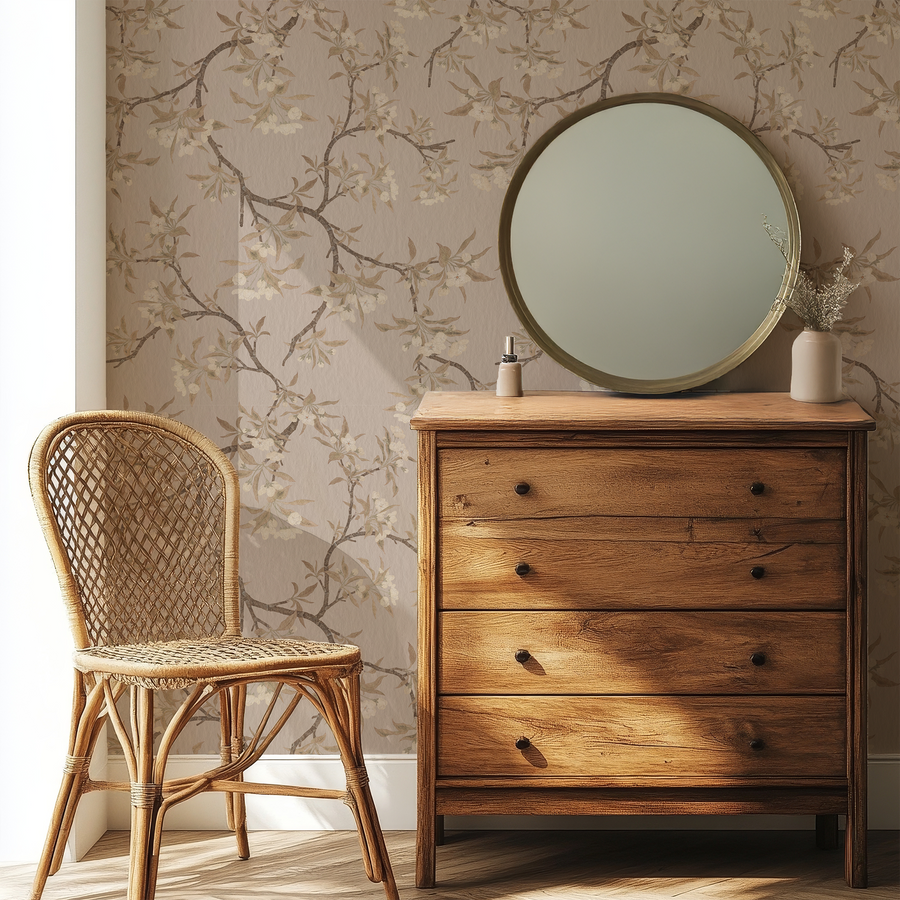 A bedroom with a rattan chair and drawers with a circlular mirror featuring 'Leah' which is traditional wallpaper featuring delicate pear blossom branches with soft pink flowers and green leaves on a subtle textured neutral background in an elegant repeating pattern.