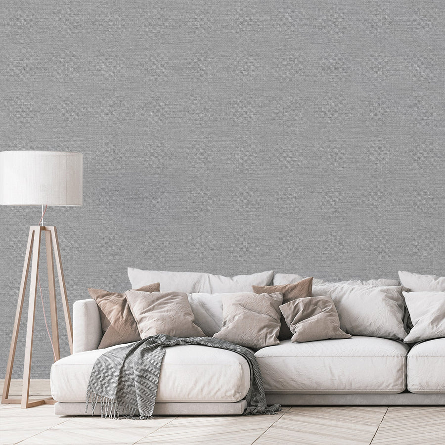 A modern living room with a white fabric sofa with pillows and a blanket draped across it, next to a standing lamp featuring grey and silver wallpaper with a woven fabric effect pattern, creating a subtle textured appearance.