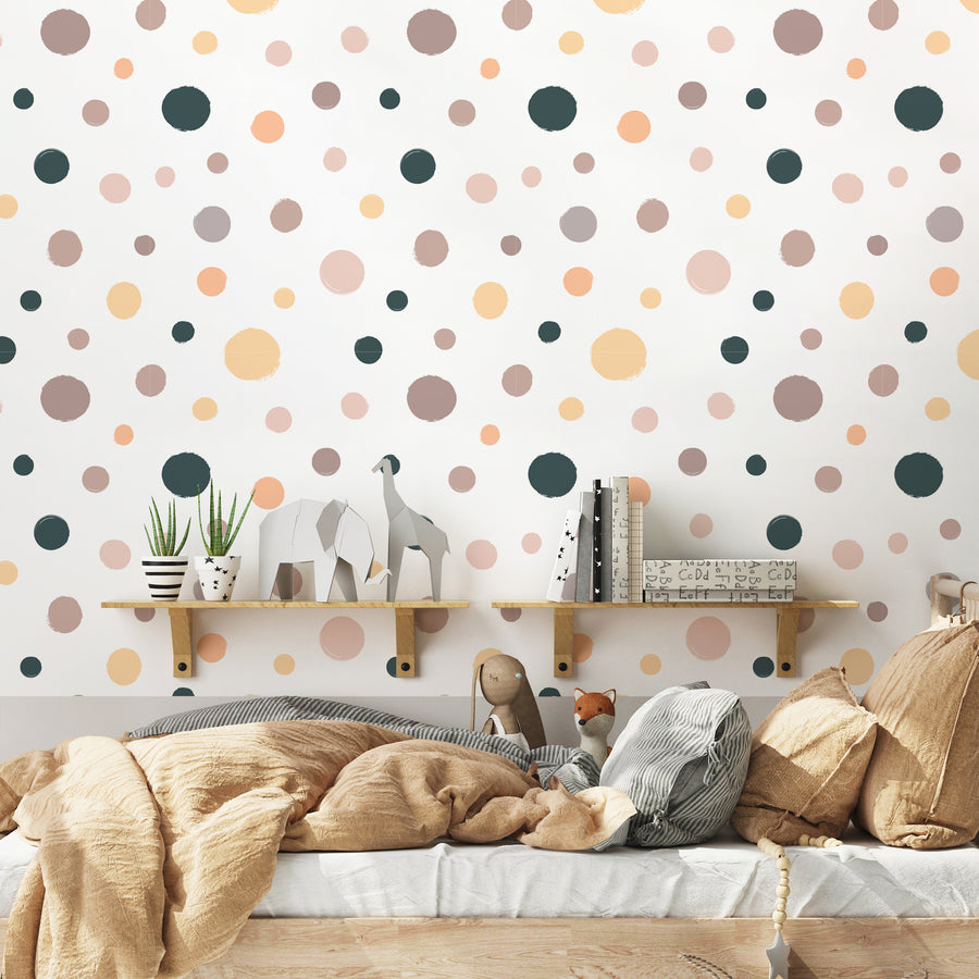 A childrens bedroom with toys, pillows and blankets on top of a bed featuring 'Isla' which is a children's wallpaper featuring multicoloured pastel polka dots in pink, yellow, purple and navy on a crisp white background in a playful repeating pattern.