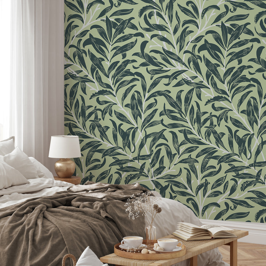 A bedroom styled in a neutral setting with bedding, lamp and a bench at the bottom of the bed featuring 'Lyra' which is a botanical wallpaper featuring layered green tropical leaves and intertwining leafy vines on a soft neutral background in a lush repeating foliage pattern.