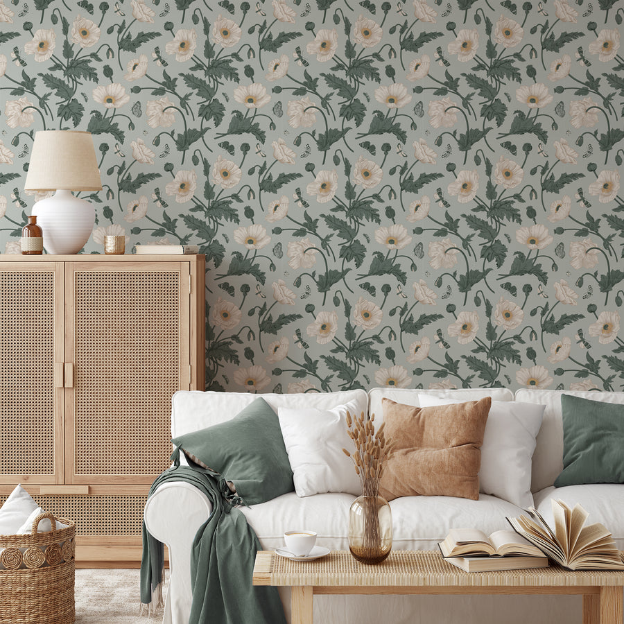 A coastal style living room with rattan furniture featuring a botanical wallpaper featuring elegant white poppies, butterflies and leafy stems on a muted sage green background.