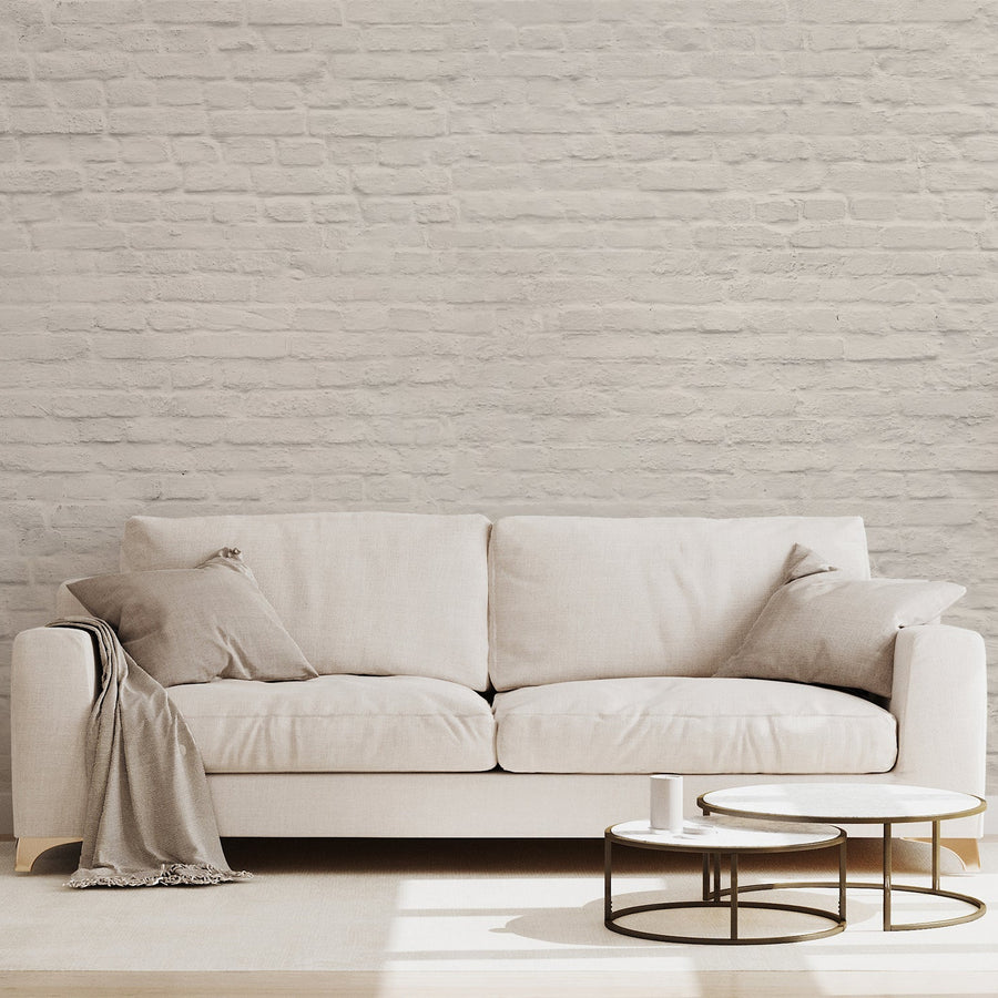 A living room with a cream fabric sofa draped with beige pillows and a blanket and has two low coffee tables showing 'Zaina' which is a cream wallpaper featuring a realistic painted brick pattern with soft neutral tones and subtle distressed texture in a classic repeating design.