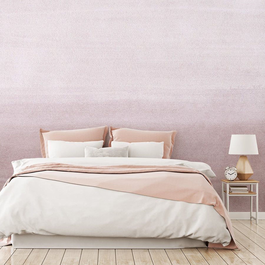 A bedroom with soft furnishings, showing 'Clara Blush' which is a blush pink wallpaper featuring a soft watercolour wash effect with a subtle ombré gradient transitioning between light and darker pink tones.