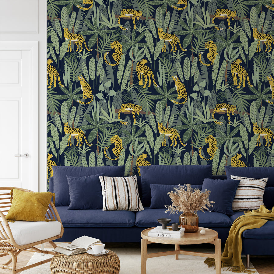 A living room with a navy sofa scattered with pillows and low coffee table featuring 'Clymene' which is a safari wallpaper featuring illustrated cheetahs among lush tropical palm leaves and exotic green foliage on a soft neutral background in a repeating jungle pattern.