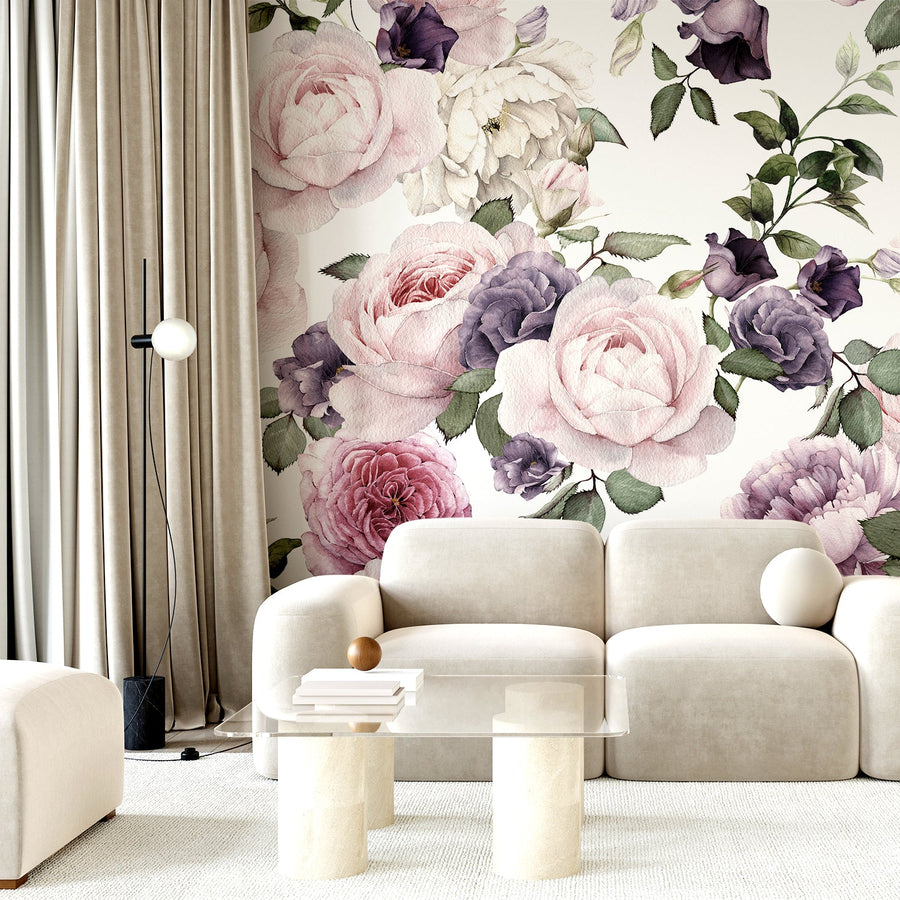 A living room showing a cream sofa with a glass coffee table and soft curtains, showing 'Rosalie' which is a pink and purple wallpaper featuring soft watercolour roses with delicate floral details in a romantic repeating botanical pattern.