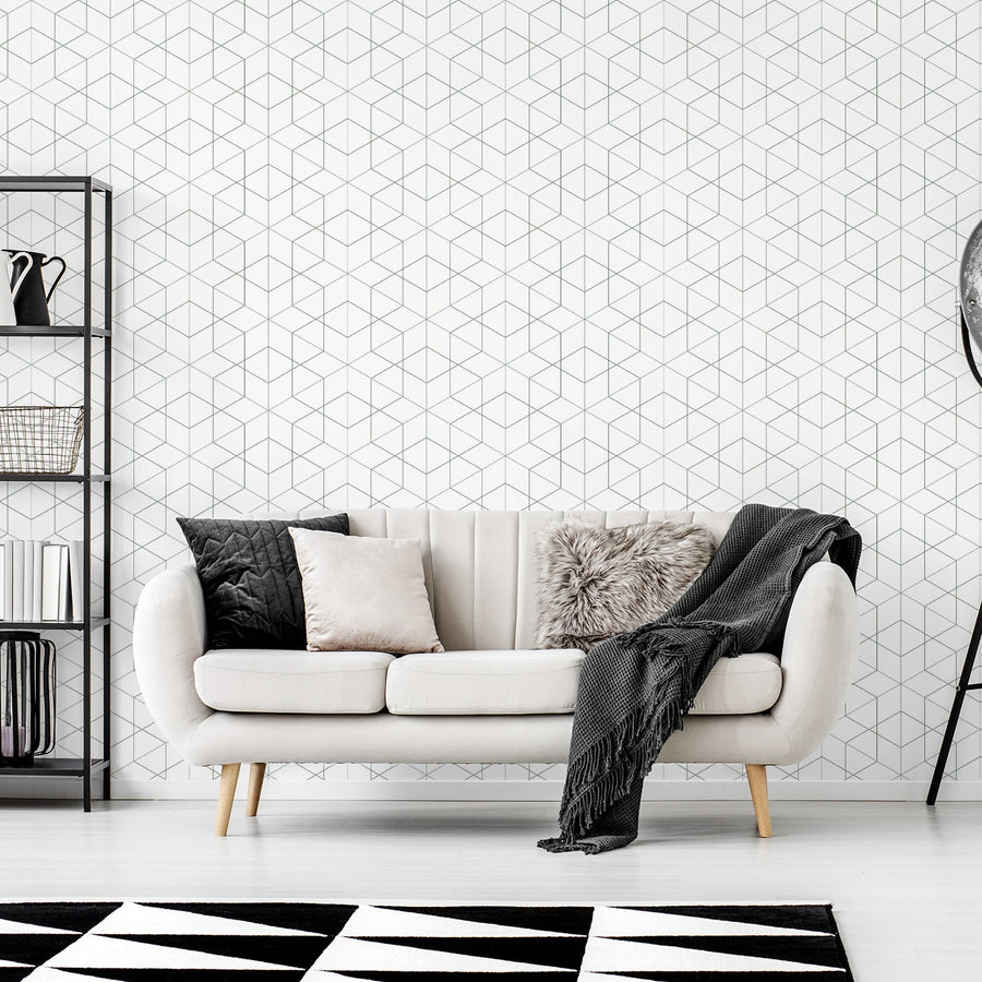 A living room with a cream two seater sofa next to a stack of black metal shelves and a free standing lamp featuring 'Rosamund' which is a dark green geometric wallpaper featuring overlapping diamond outlines and fine linear detailing in a modern repeating pattern.